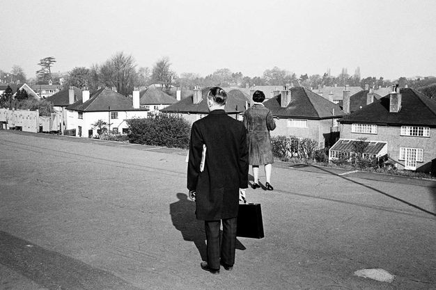 Black and white photograph. A man and a woman stand along a street, facing away from us. The man wears a black suit and coat has a briefcase in one hand and a newspaper tucked under the other arm. Light reflects off his dark hair. The woman wears a knee-length coat over bare legs and low heels. A sea of identical rooftops fills the view beyond.