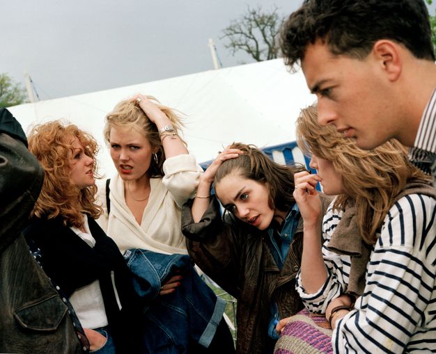 Colour photograph. The heads and shoulders of five pale-skinned young men and women nearly fill this picture. The two women at the centre run their hands through their hair in the same gesture. Two other women have feathered hairstyles. A tent fills much of the background behind the group.