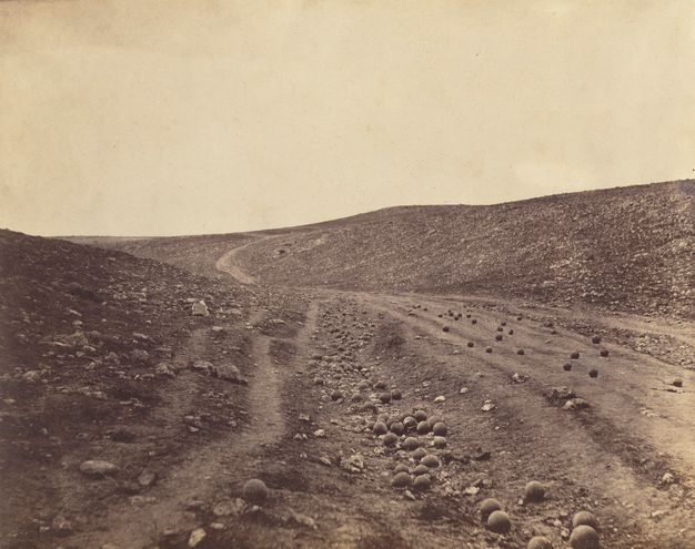 Black and white photograph. Dozens of cannonballs are strewn along a road and ditch in a shallow, barren valley.