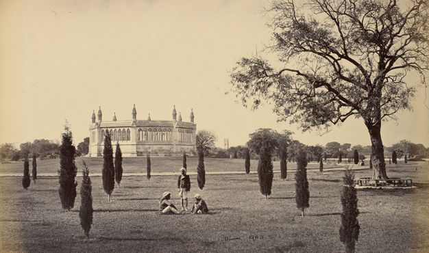 Sepia-toned photograph. Two men sit and one stands in a grassy park dotted with thin trees. The men have dark skin and wear light-coloured turbans. Across the field is an octagonal, temple-like structure.
