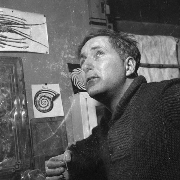 Black and white photograph. Shown from the chest up, a man’s body angles to our left as he looks sharply up under raised brows. Smoke wafts from a thin cigarette pinched between thumb and forefinger, along the bottom edge of the image. Works of art hang on the wall behind him.